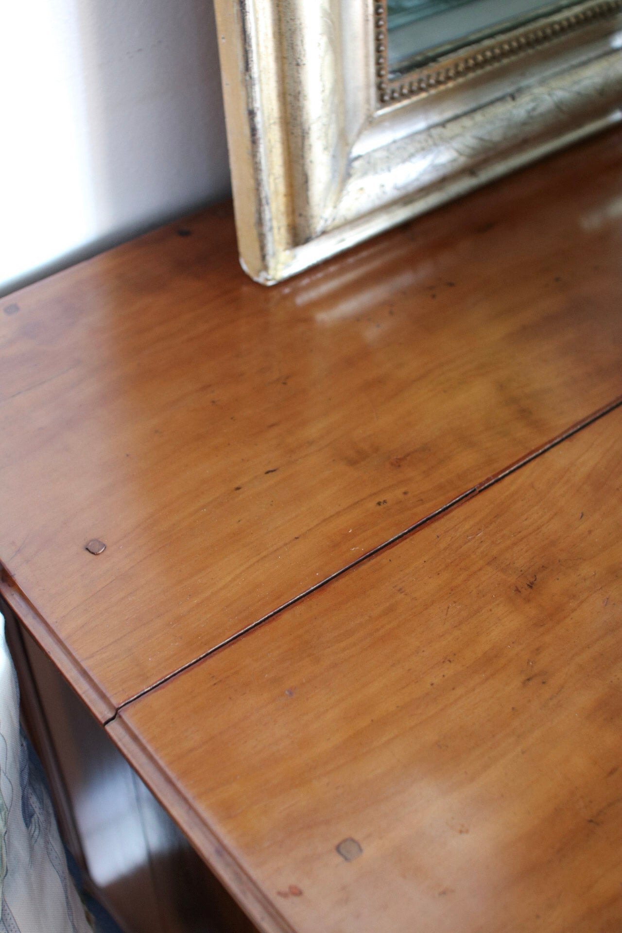 Close-up of a warm brown wooden table surface with visible grain, a seam line, and part of a distressed gold-framed mirror resting on top.