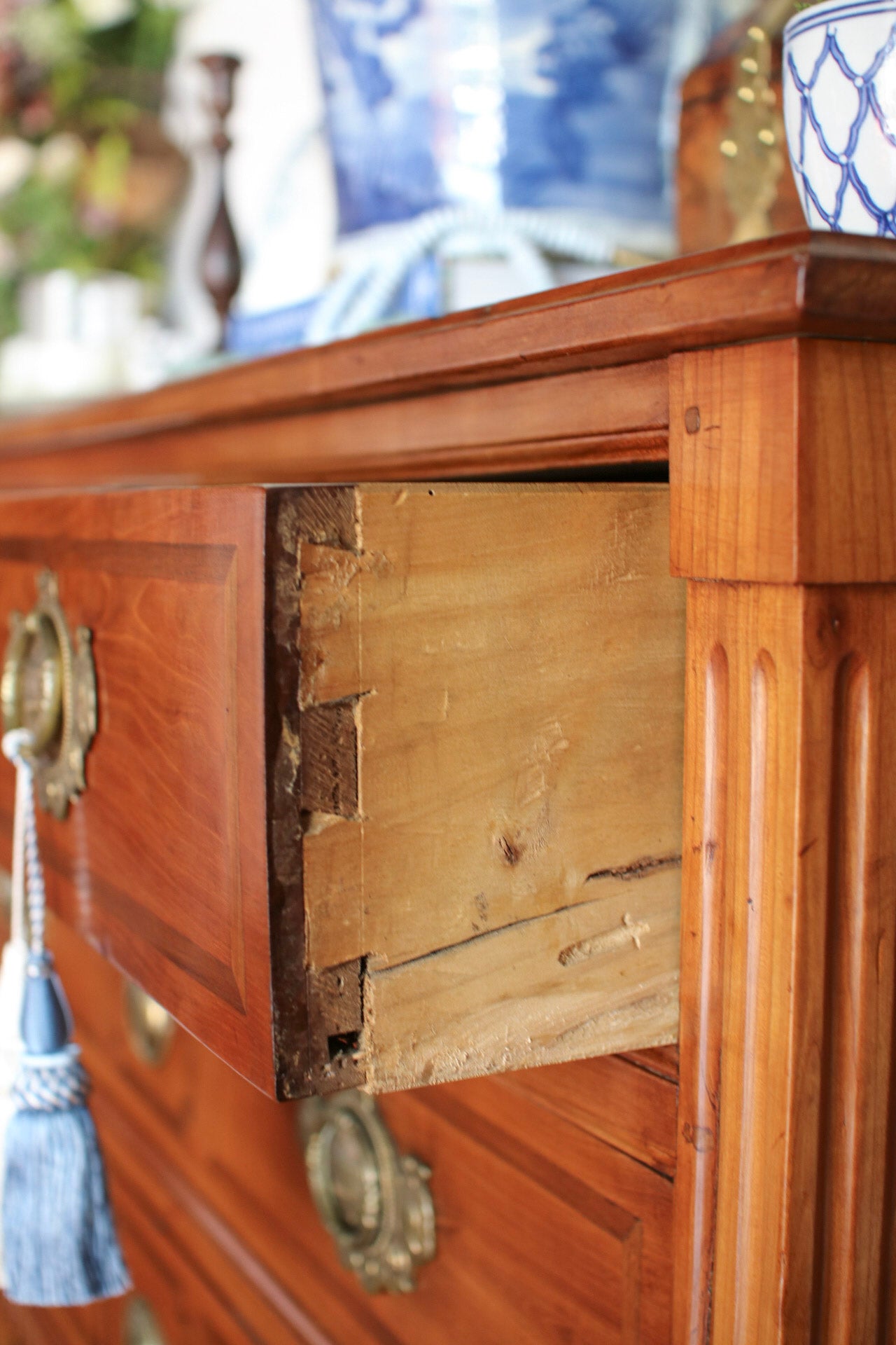 Close-up of a warm medium-brown wooden dresser with a drawer pulled open to reveal the interior joinery, featuring brass-style hardware and a blue tassel accent.