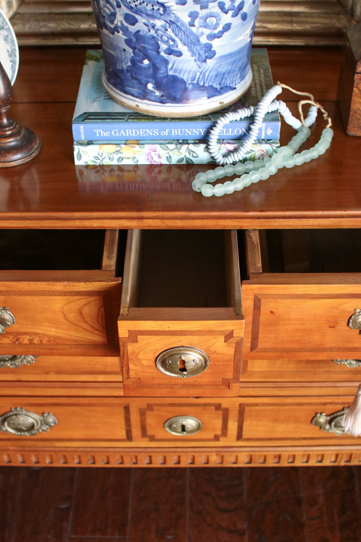 A close-up of a polished wooden cabinet or desk with multiple drawers, one open. A blue-and-white ceramic pot sits on top of two books, next to a beaded necklace.