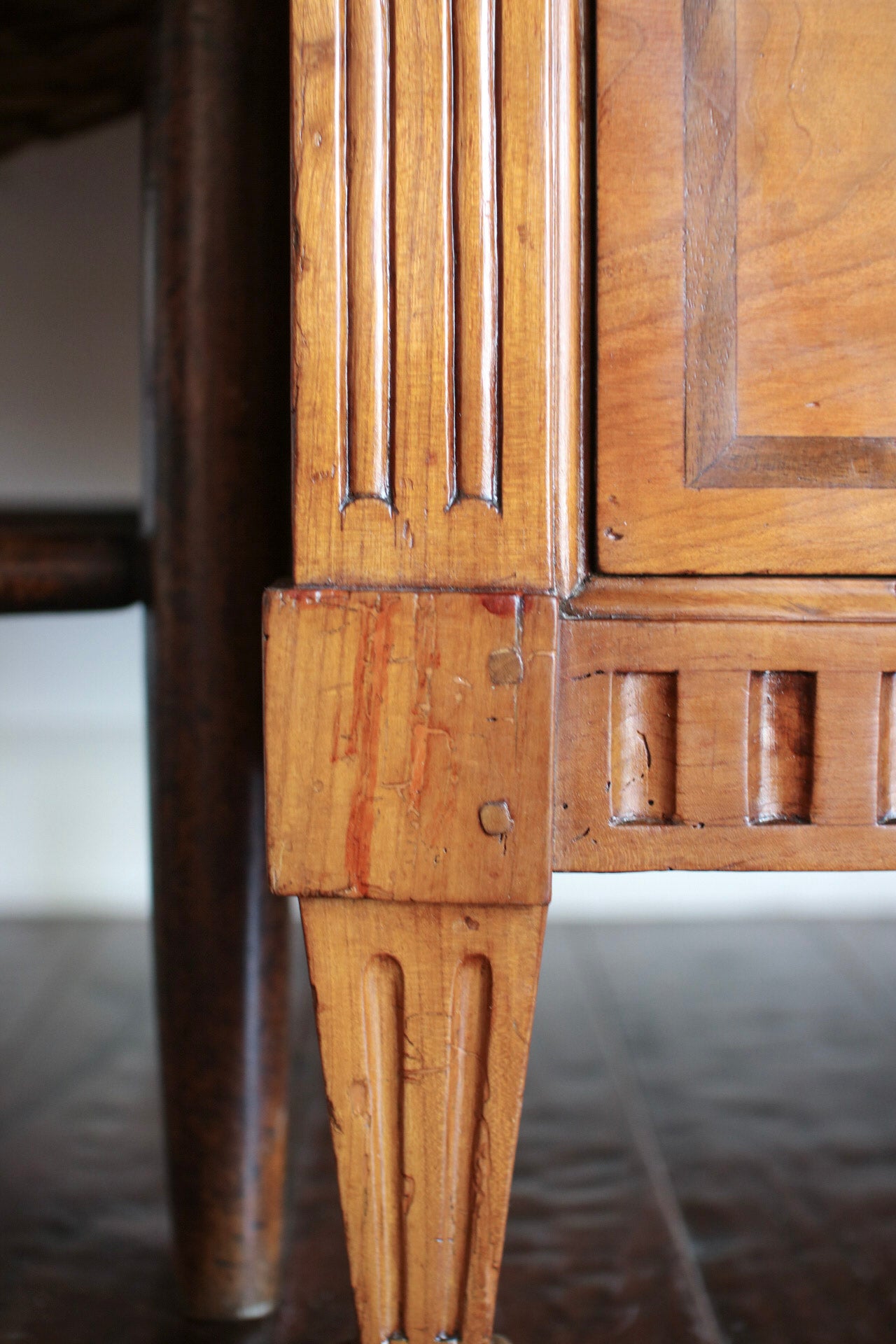 Close-up of a wooden cabinet leg with decorative fluted detailing and visible wear and scratches on the wood finish.