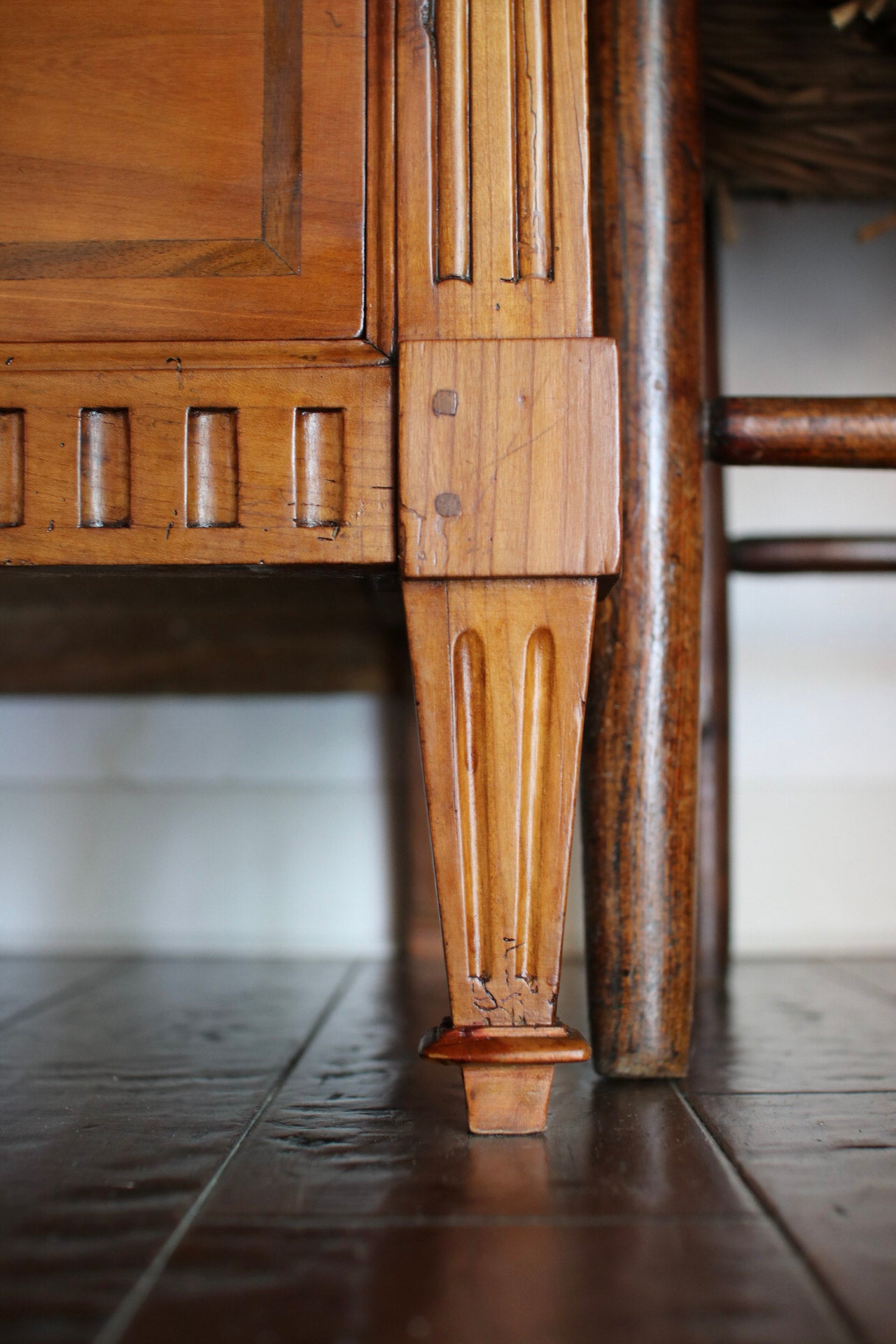 Close-up detail of an antique wooden piece of furniture, showing a carved wooden leg foot and decorative joint on a dark wood floor.