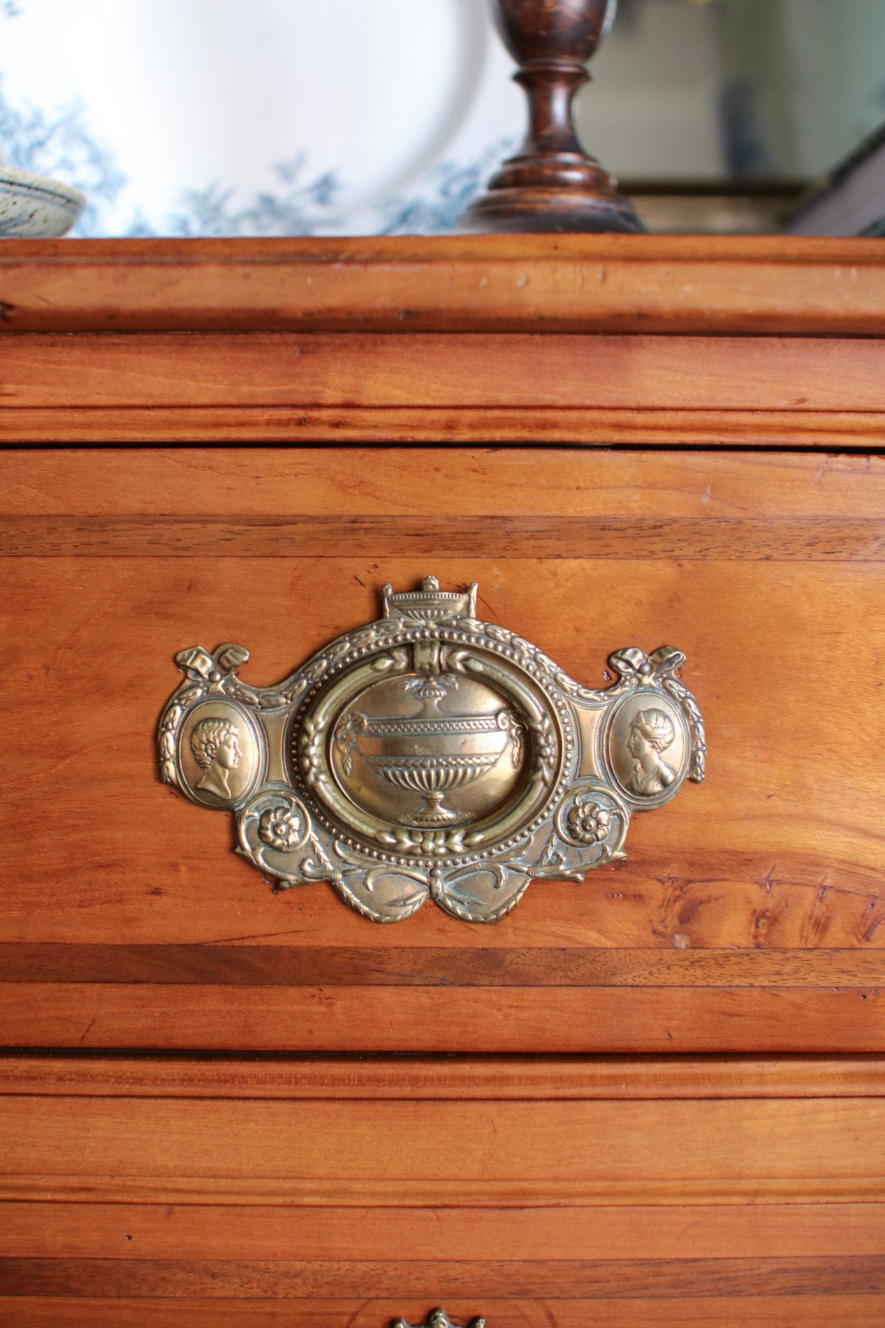 Close-up of a vintage ornate brass drawer pull handle on a warm brown wooden chest or cabinet.